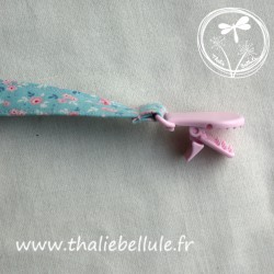 Attache tétine bleu à fleurs roses en tissu coton, avec une pince plastique rose qui s'utilise facilement